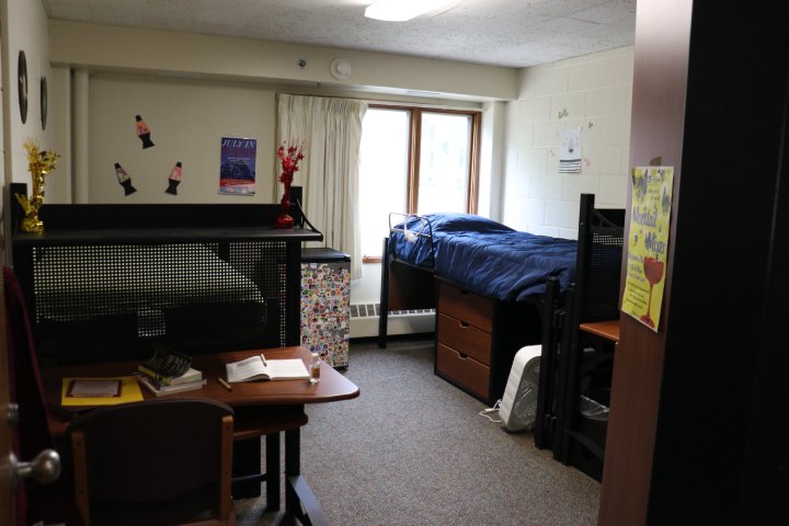 A dorm room with a bed on the right side, dressed with a blue comforter and placed over wooden storage drawers. The window behind the bed is covered with white curtains, and a small fan is on the floor near the bed. On the left side of the room is a desk with books and a chair.